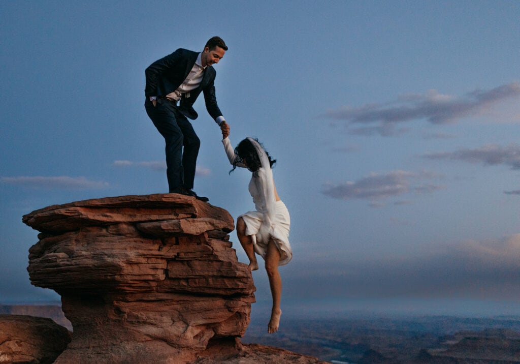 A groom helps his bride onto a ledge at Dead Horse Point at Sunset