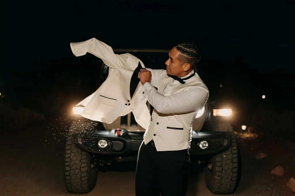 The groom puts on his suit jacket outside in front of the jeep headlights.