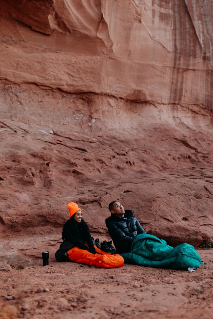 A couple sits in their sleeping bags watching the view as the sun starts to rise. 