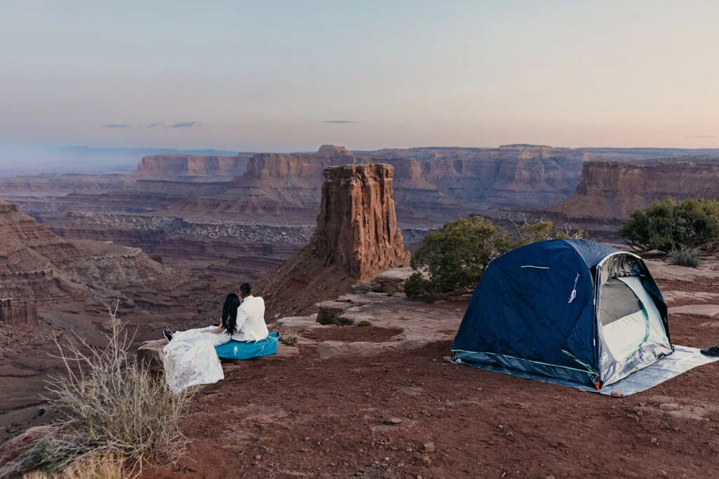 A couple sits on the edge of a cliff watching sunset on a vista by there tent.