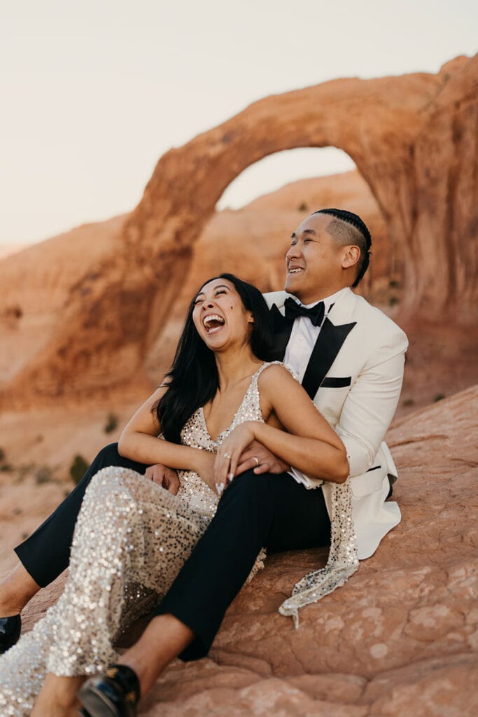 The couple sits together in the desert laughing in beautiful attire. 