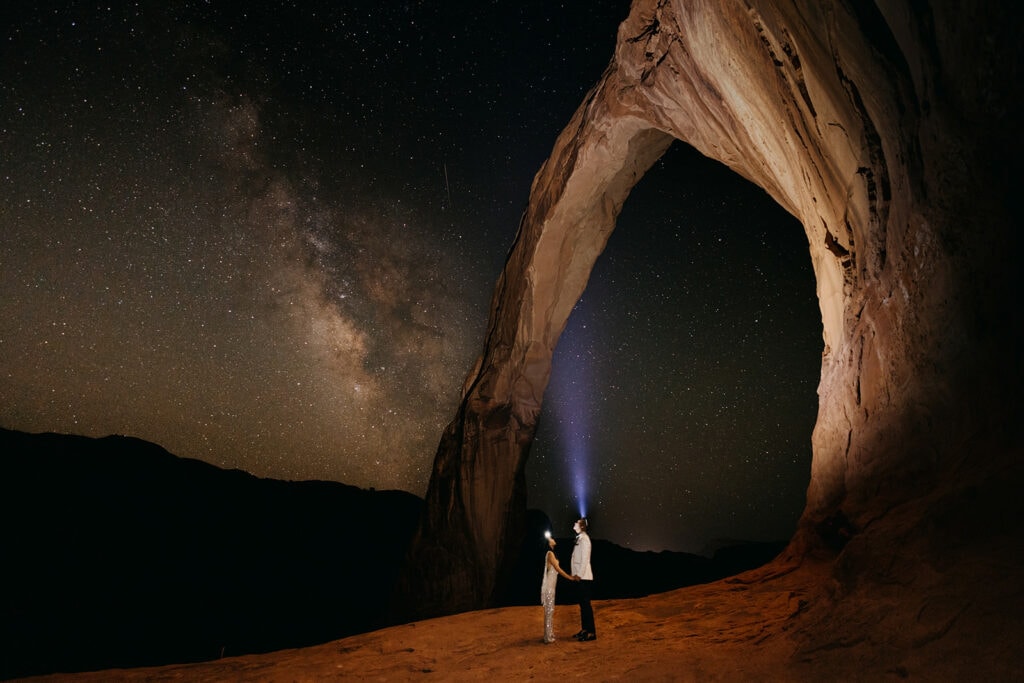 A couple stands under an arch in the night with their headlamps on as the milky way shines above. 