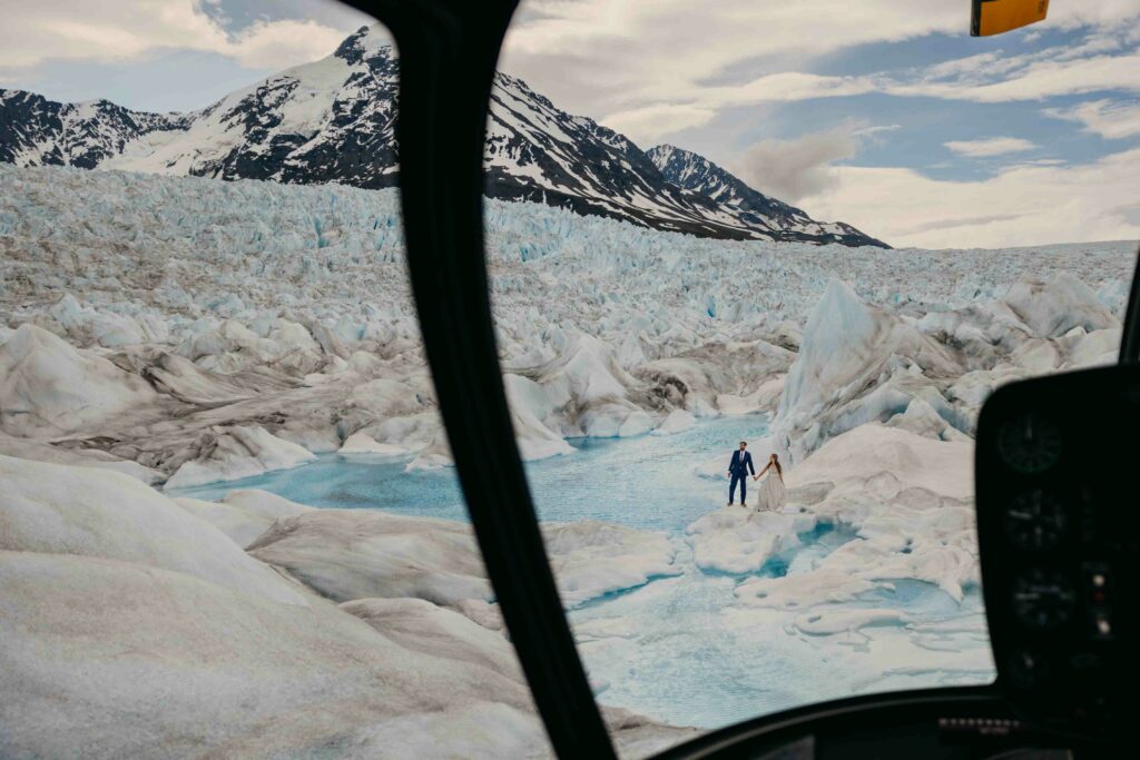 A couple walks on a glacier as a photo is taken from inside of a helicopter.