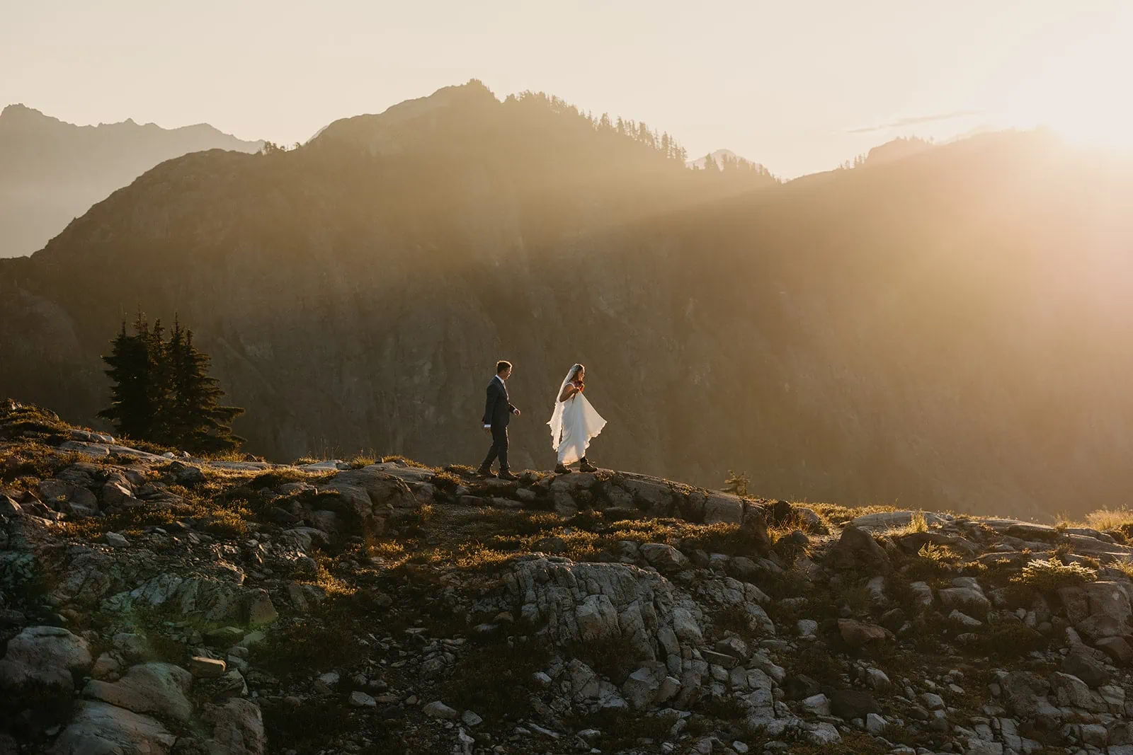 A bride and groom walk down the side of a mountain in the morning light.