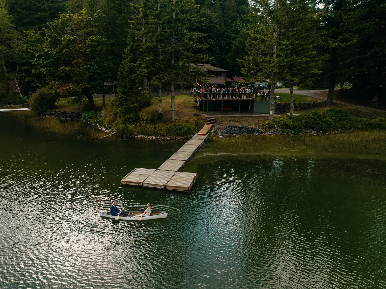 a bride and groom canoe on a lake together while their guests look down on them from a deck
