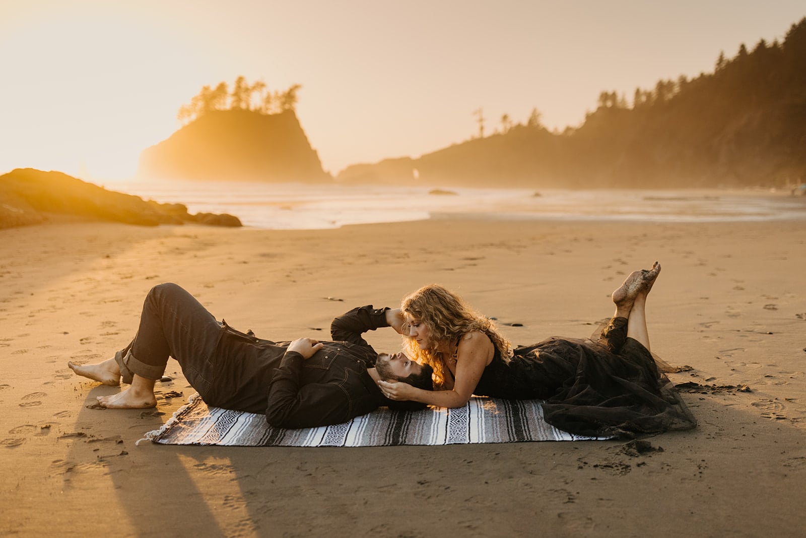 A couple lays together on a blanket on the beach at sunset. 