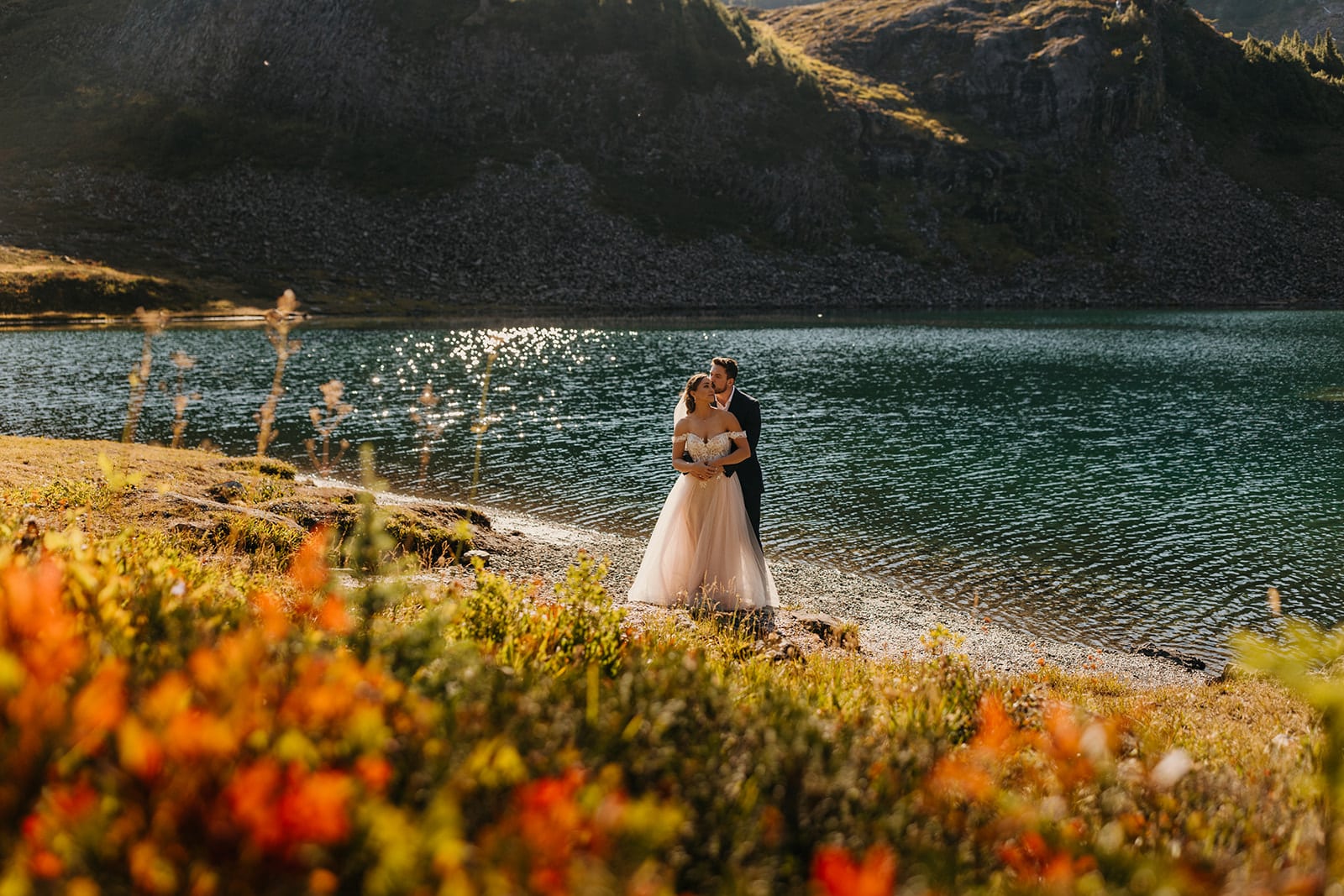 A couple has their portrait taken by a lake in the mountains on a sunny fall day. 