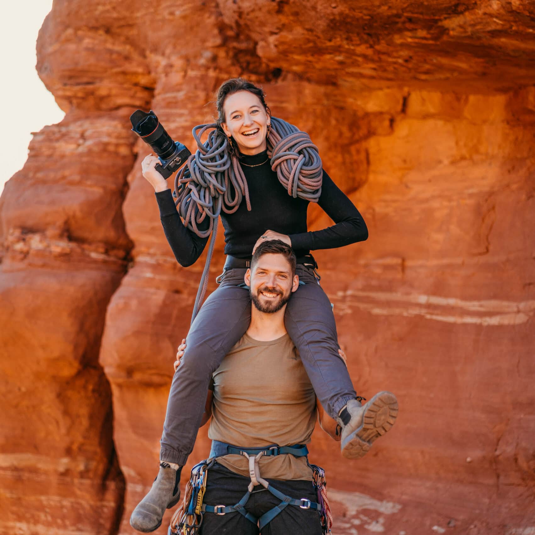 A man holds a woman on his shoulders while they both have climbing gear and ropes on them. The woman is holding a camera and they are both smiling.