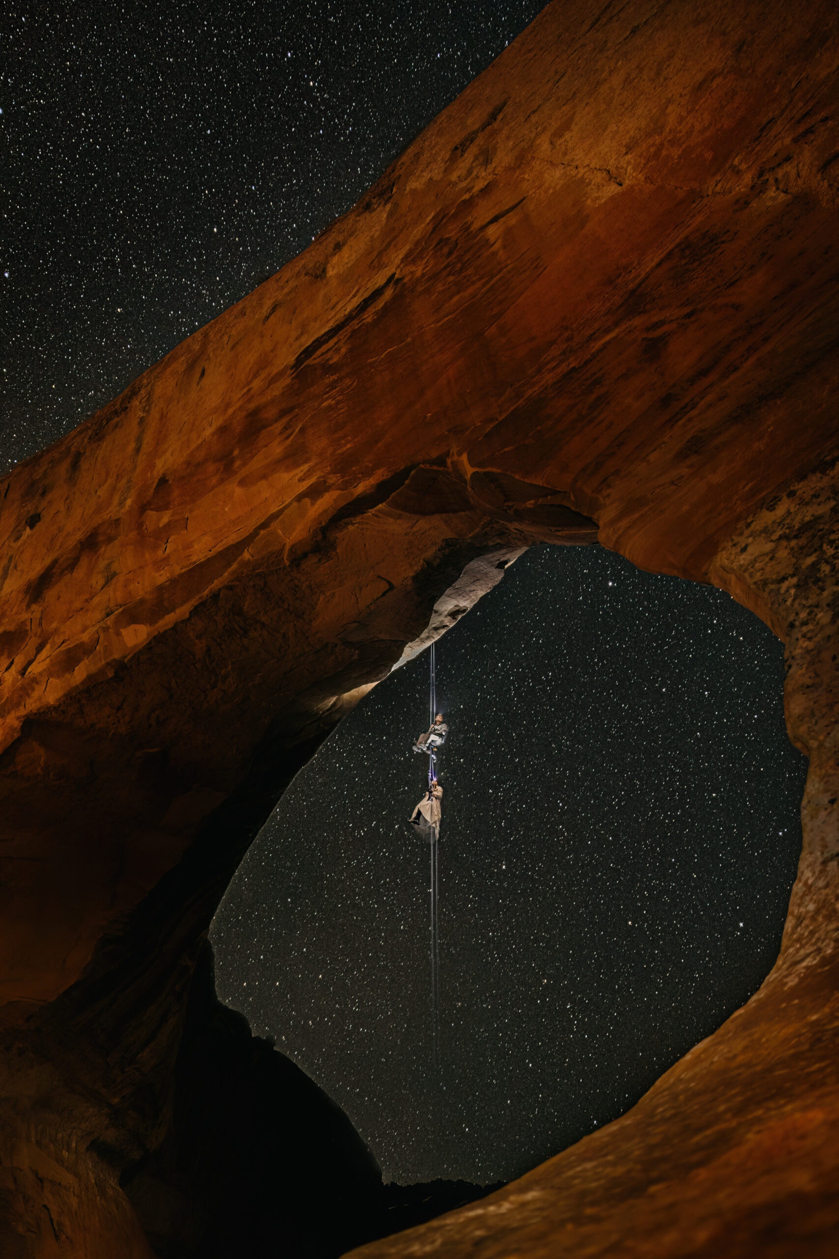 A couple rock climbs at night on sandstone in their wedding attire with the stars around them.