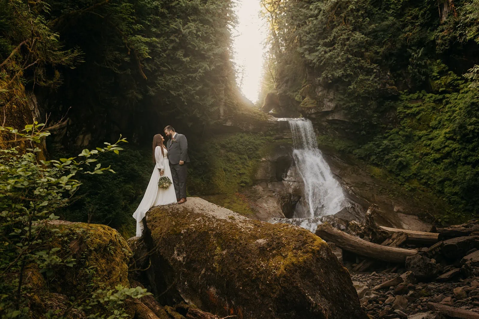 A couple holds each other close near a waterfall in the forest.