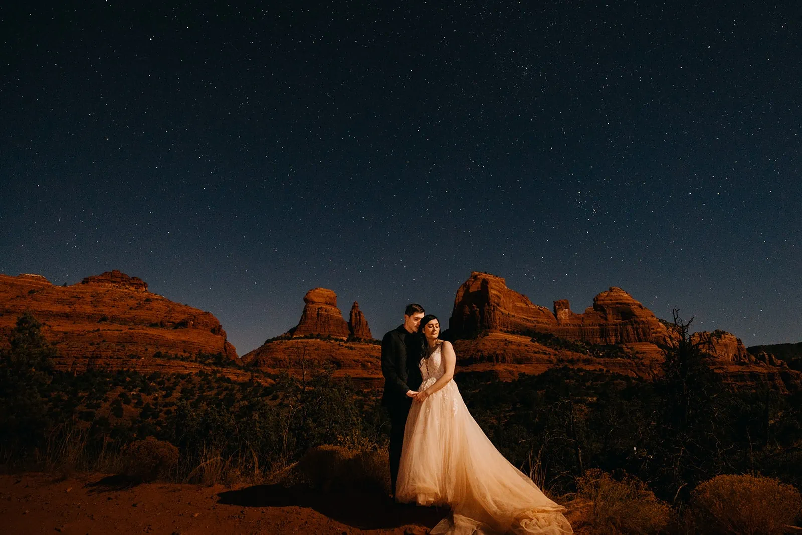 A couple stands together under the moon light and stars with red rocks behind them.