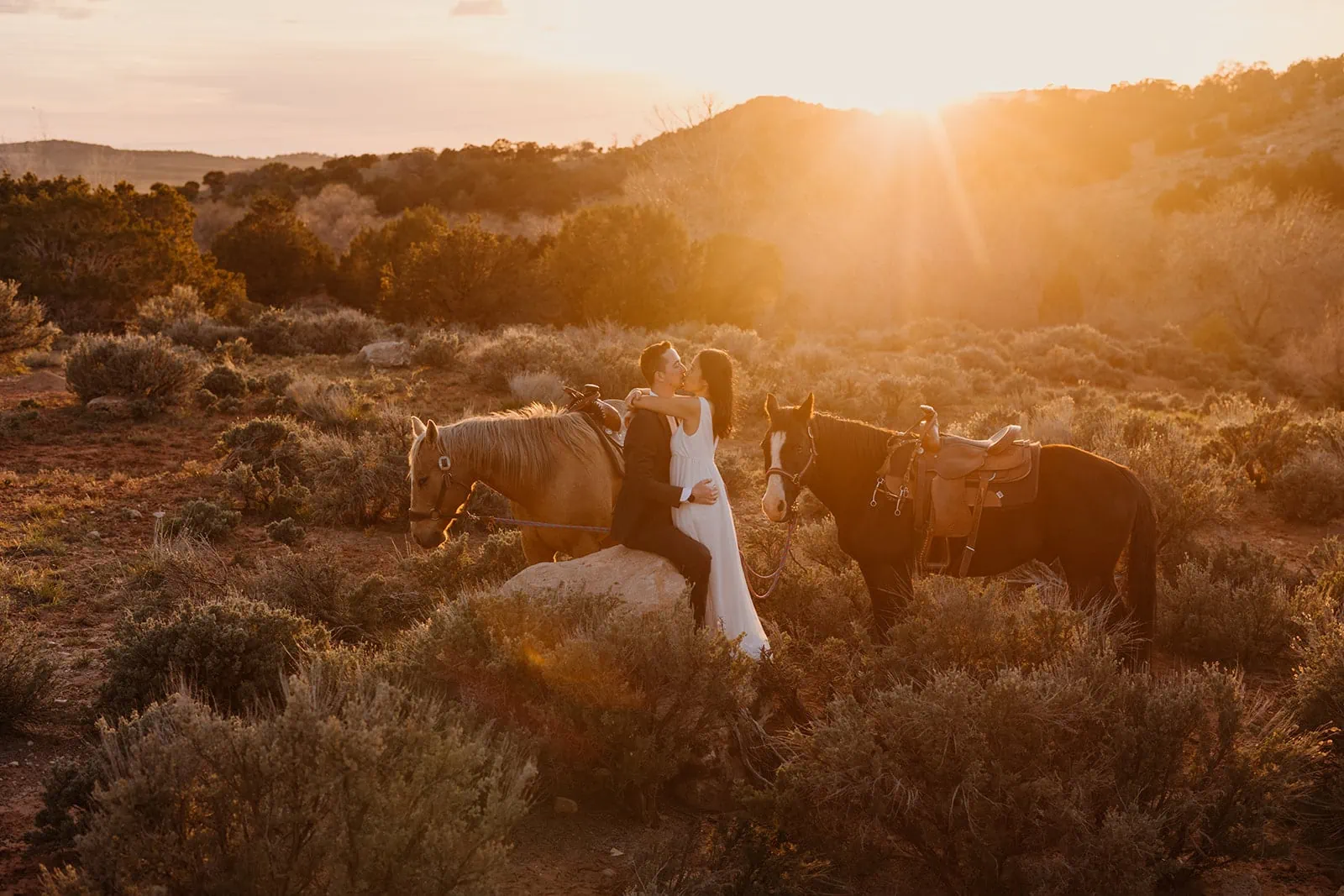 A couple holds each other romantically as the sunset and their horses stand beside them.