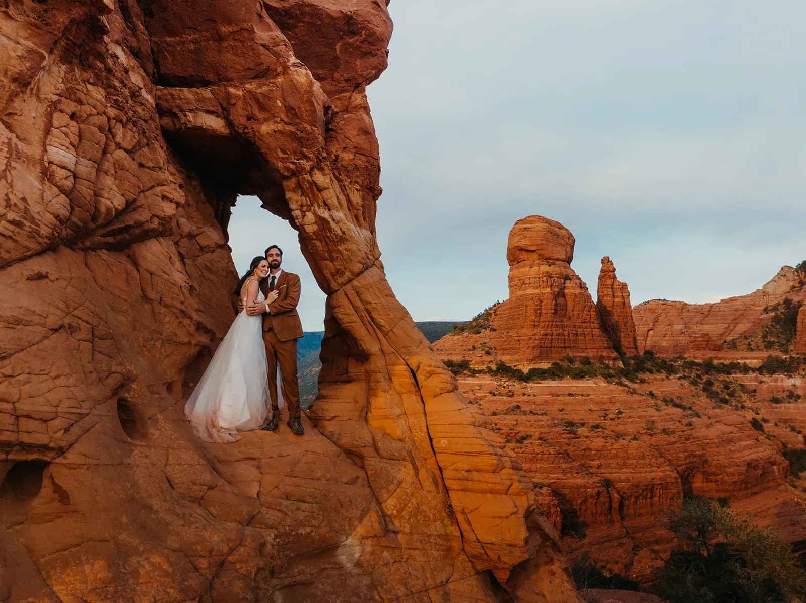 A couple stands in an arch way of red rock looking out at the view.