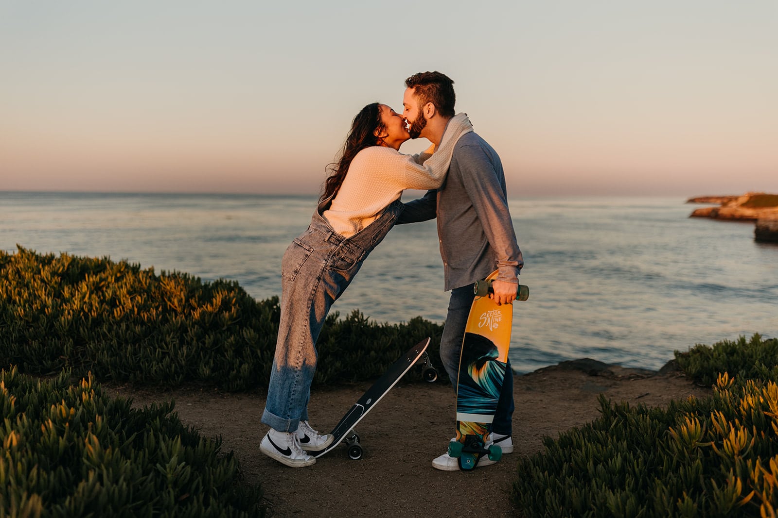 A couple shares a kiss together while skateboarding around the coast.