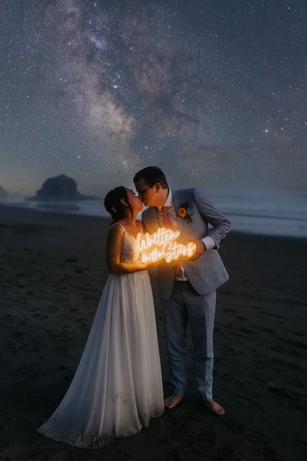 A couple holds a sign that reads written in the stars as they kiss under the milky way sky.