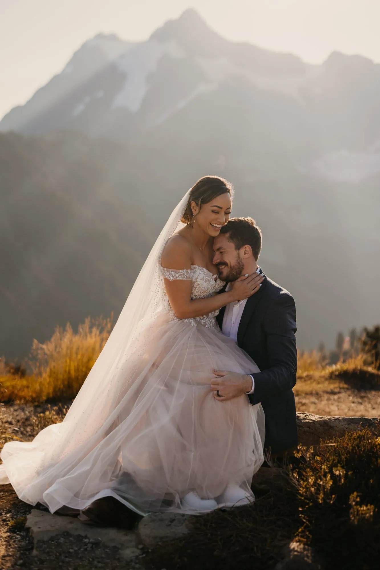 A bride and groom sit together laughing in the morning light in the mountains. 