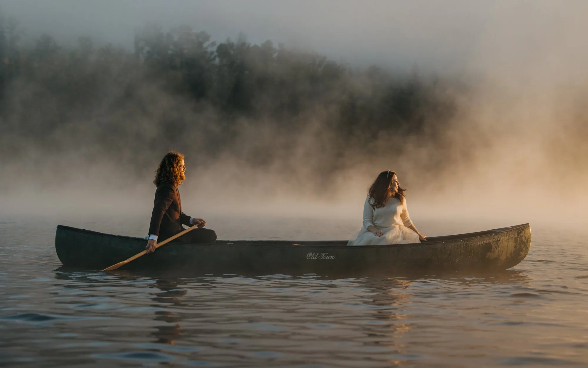 A bride and groom sit in a canoe and look at the morning light through the fog.