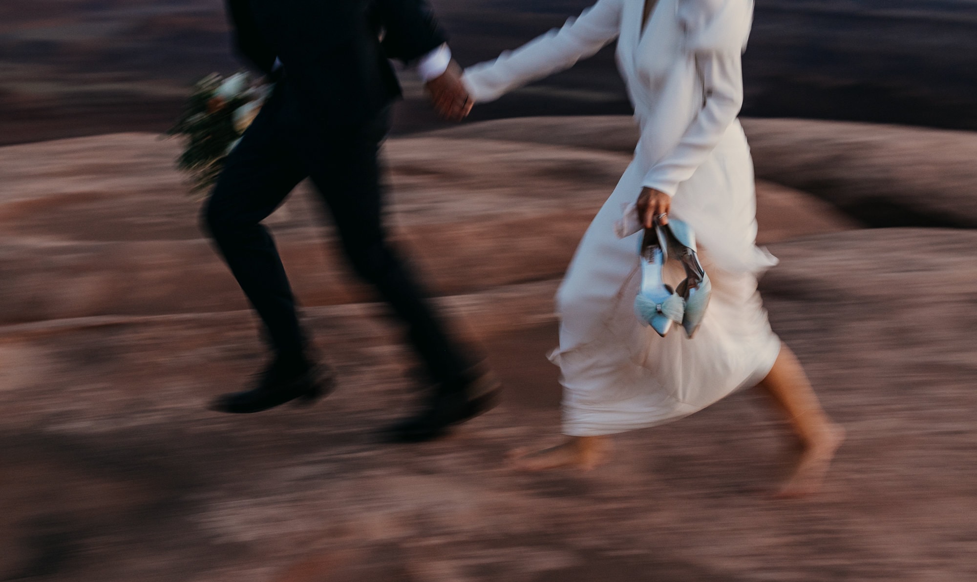 A bride and groom hold hands and run as the bride holds her blue shoes.