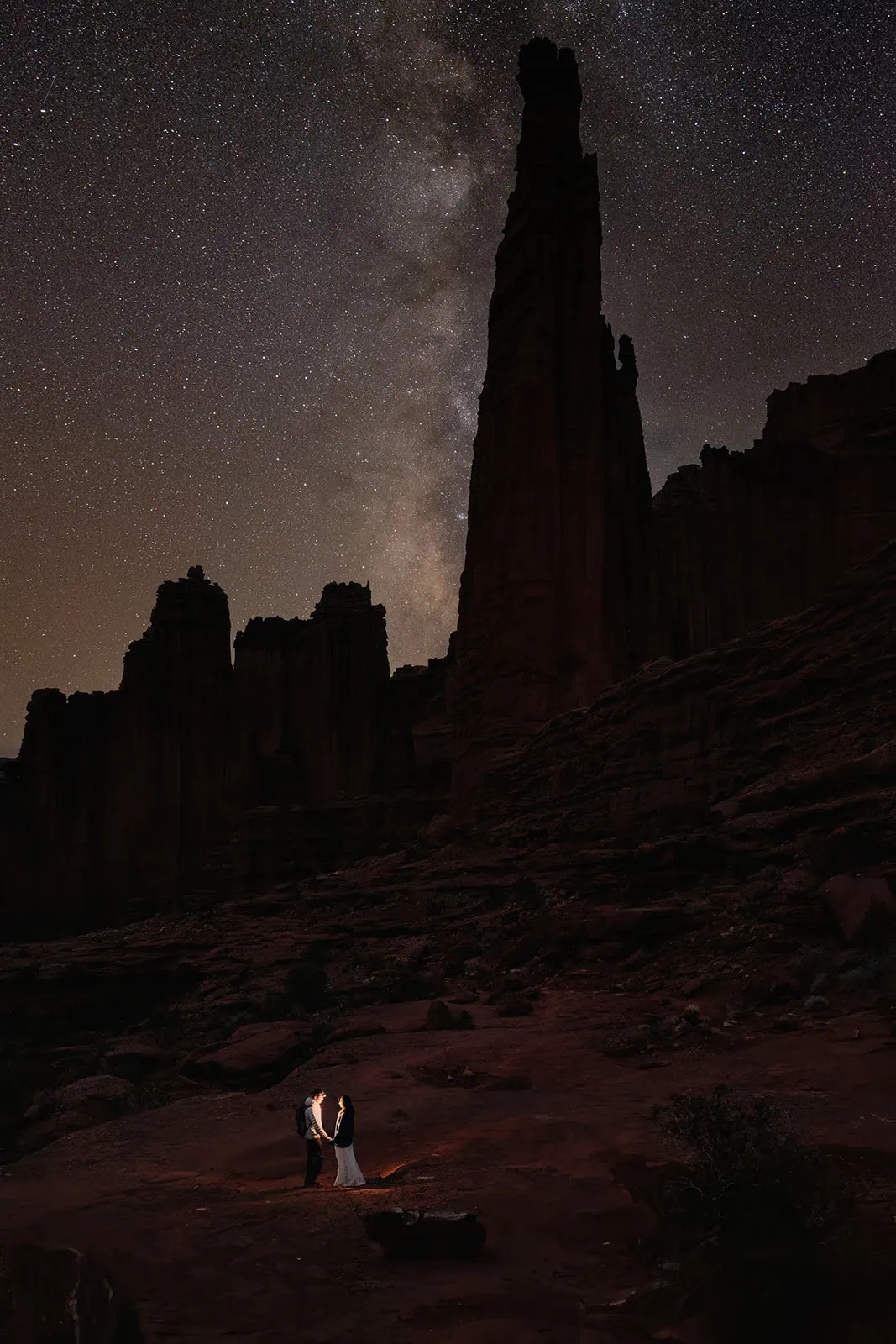 A couple stands together at night with the milky way in the sky at Fisher Towers
