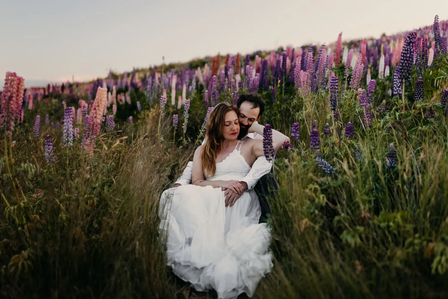 A couple cuddles up with each other as they sit in a lupine field.