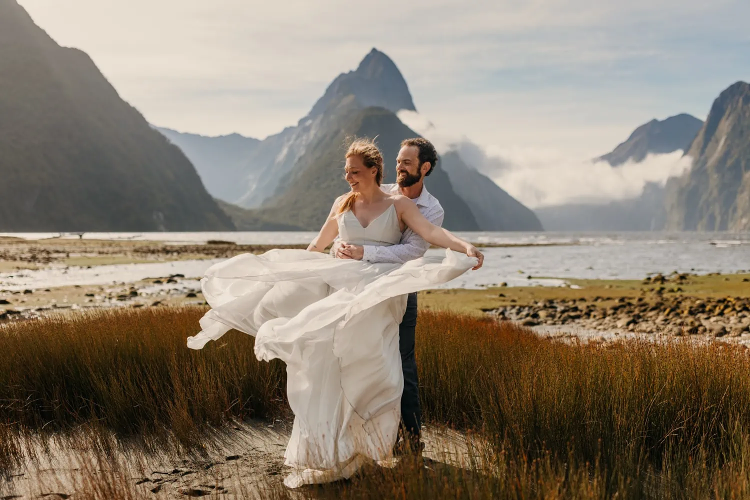 A bride lets the wind catch her dress on a sunny day at Milford Sound while her husband holds her.