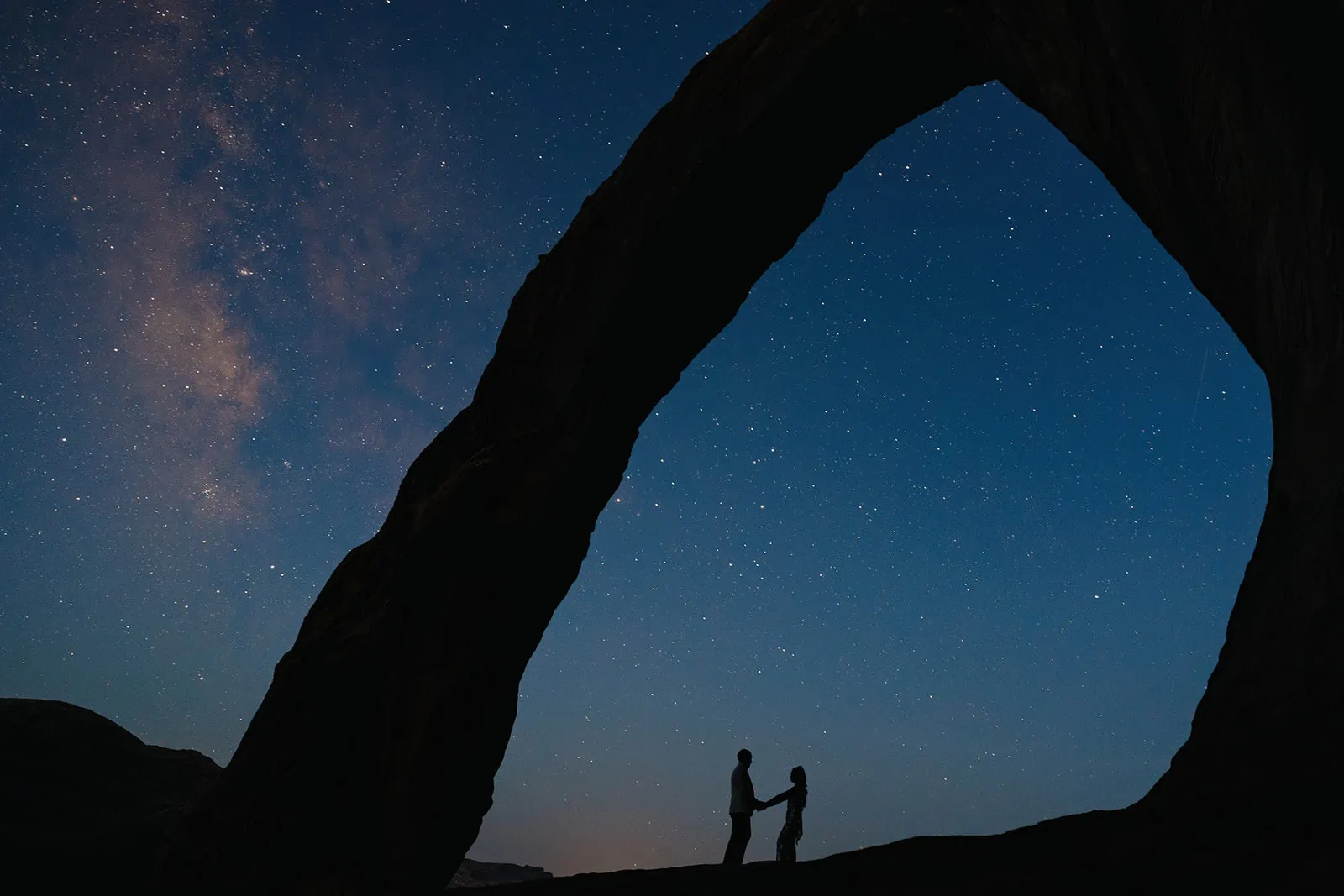 A couple stands together with an arch overhead and the milkyway in the background