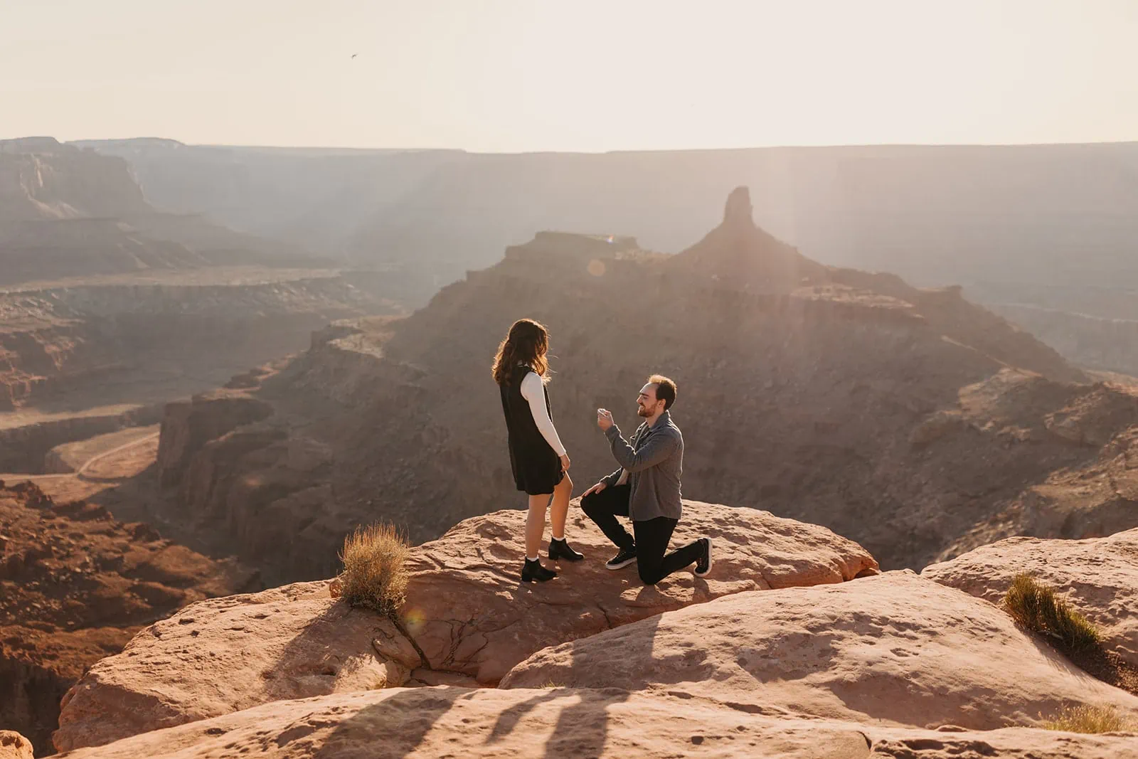 A man gets down on one knee to propose to a woman at a high up vista point in Moab