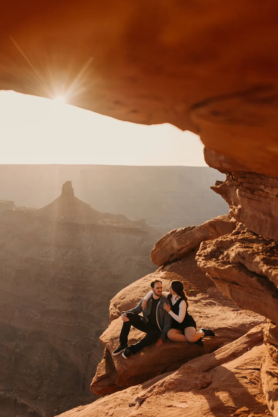 A man and woman sit together on a high up ledge at sunset in Moab