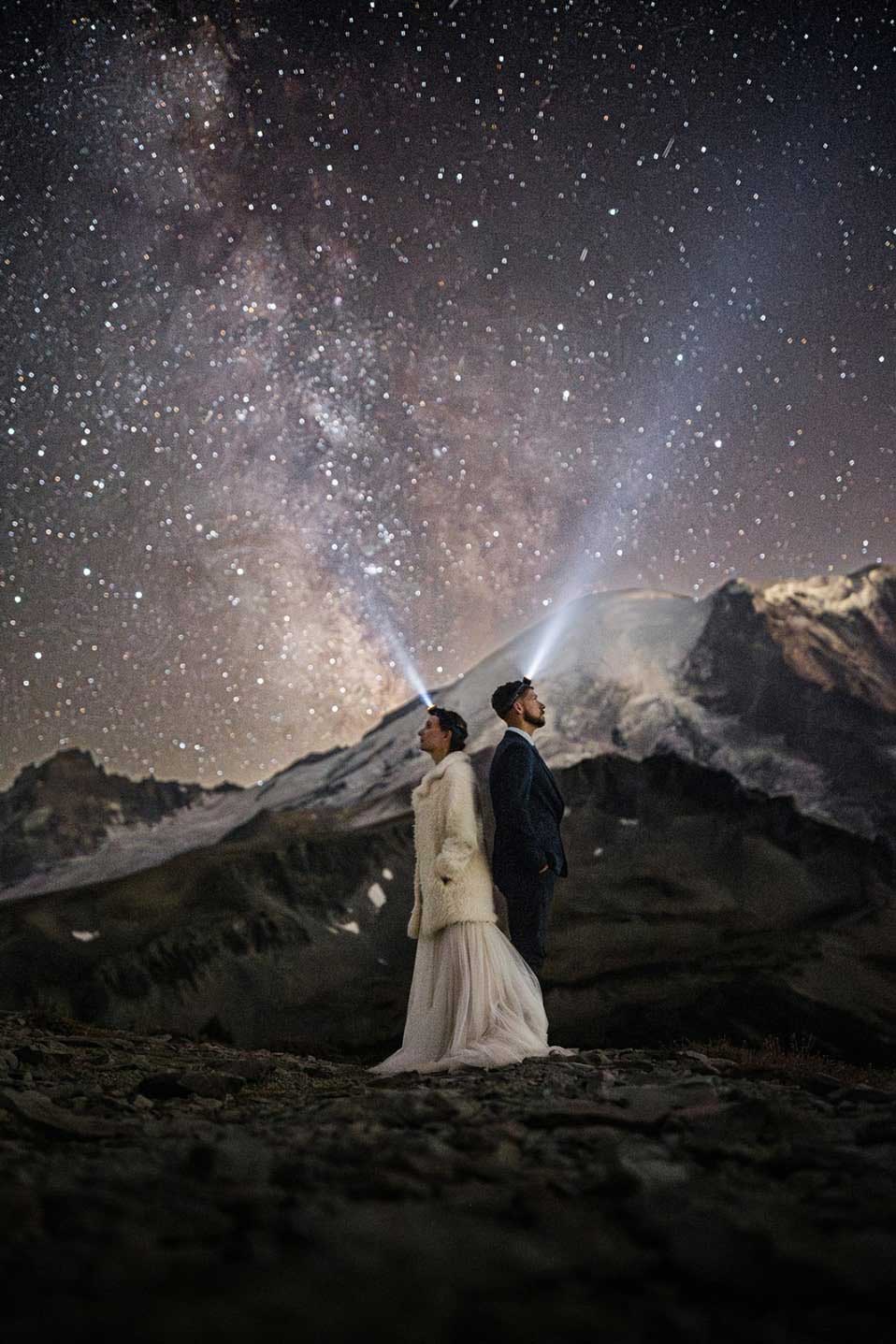 a bride and groom stand together with the milky way behind them and mount rainier with their headlamps on.