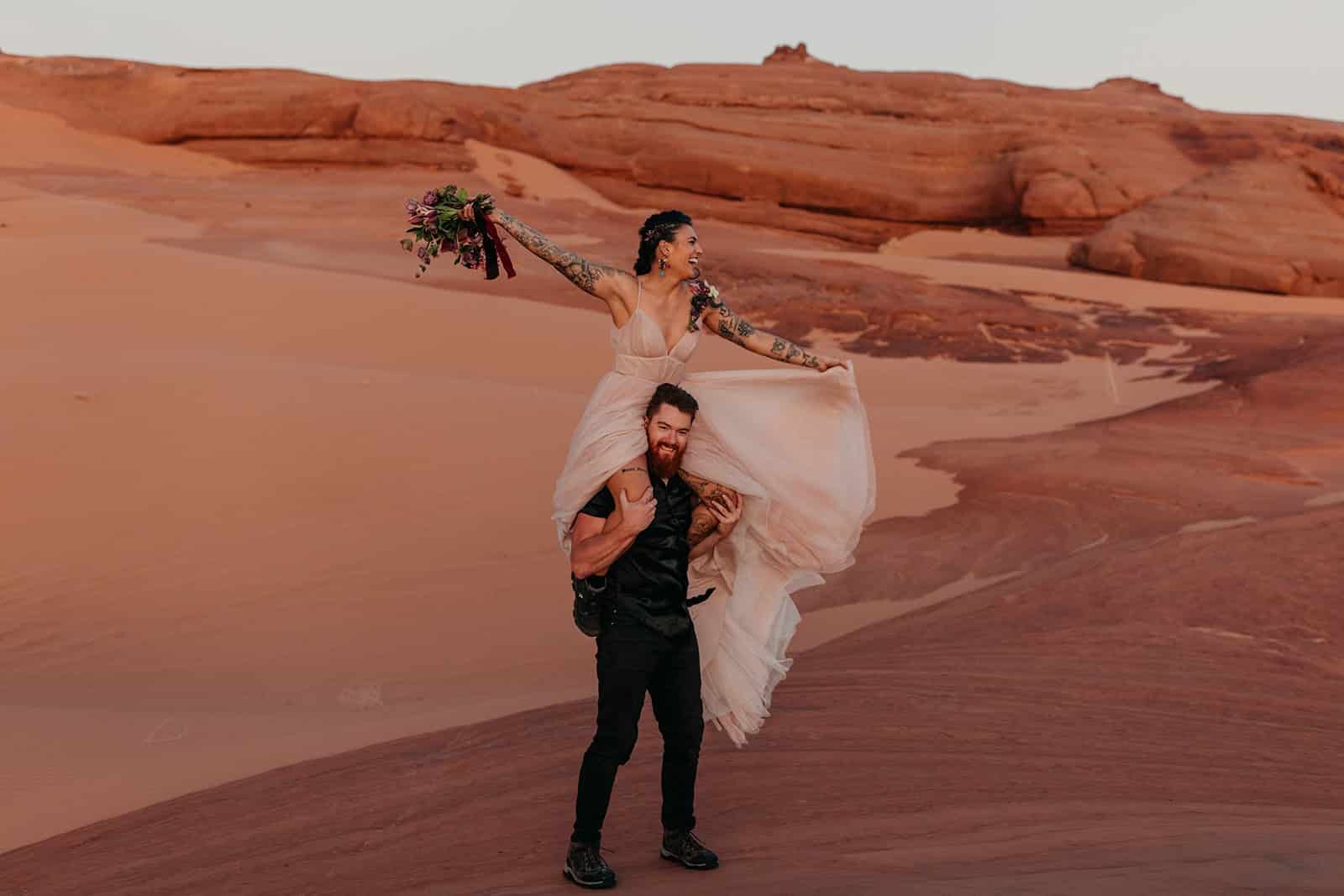 A bride is on a grooms shoulders at the sand dunes for a playful photo.