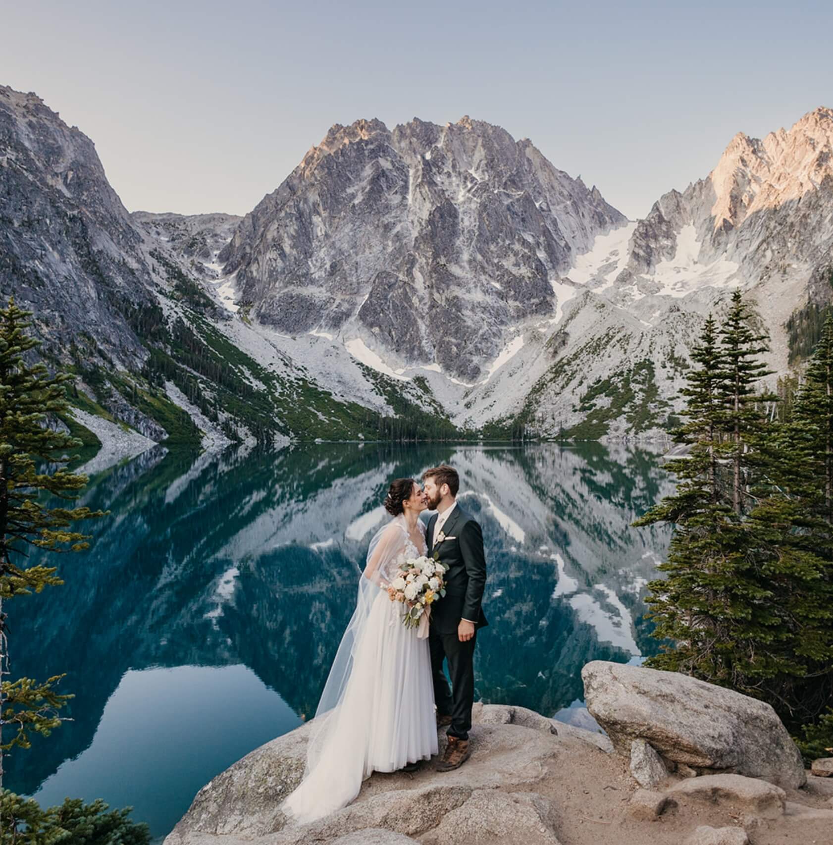 A wedding couple kissing while standing in front of a lake in the mountains