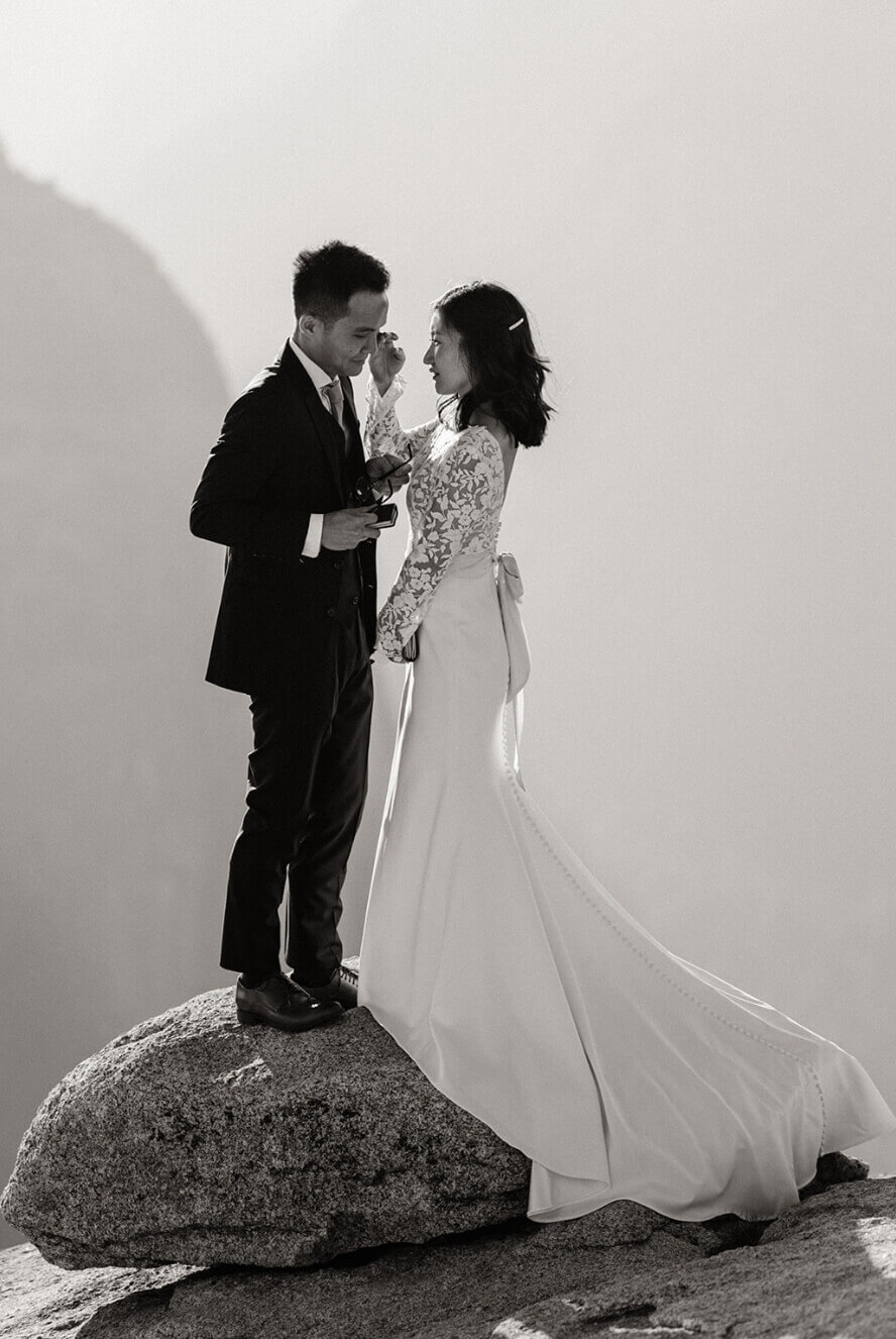 A person wiping their partner's tears as they stand on a rock in wedding outfits.