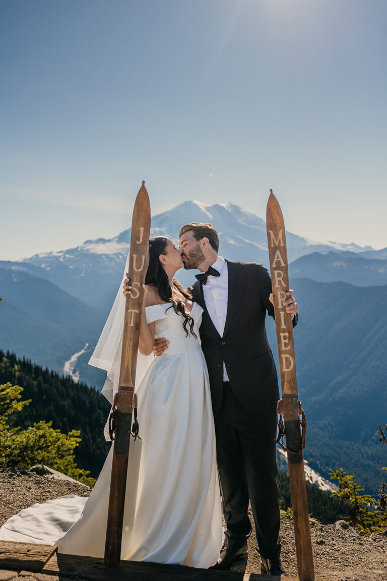 A bride and groom stand together holding skis that say just married with Mt Rainier in the background