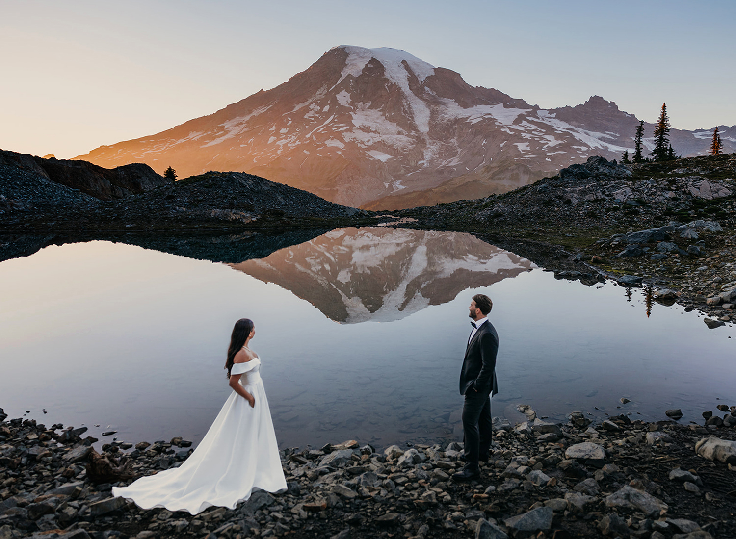 A bride and groom stand together looking towards Mt. Rainier at sunset, framed by an alpine lake.