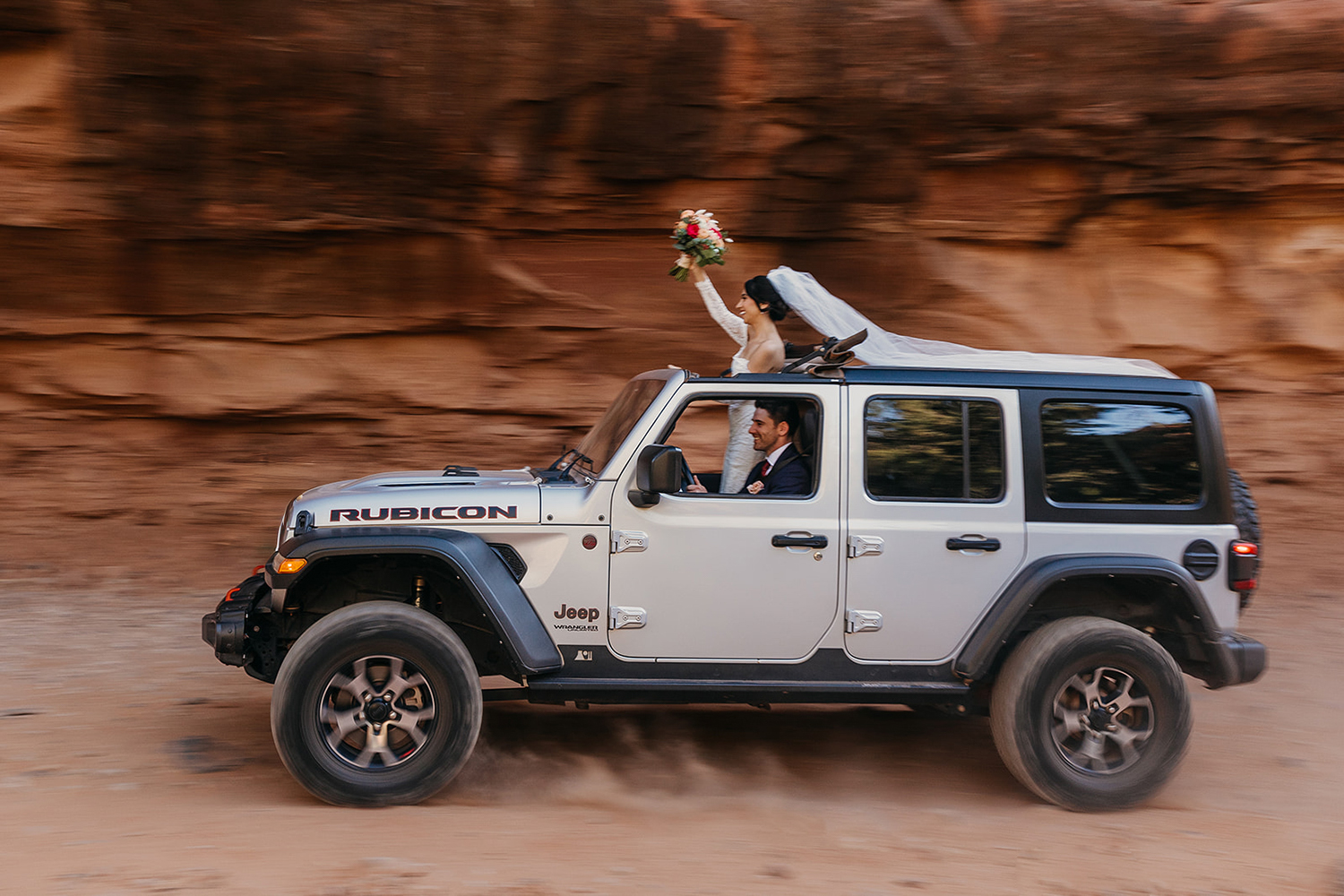 A bride standing out of the roof of her jeep while the groom drives.