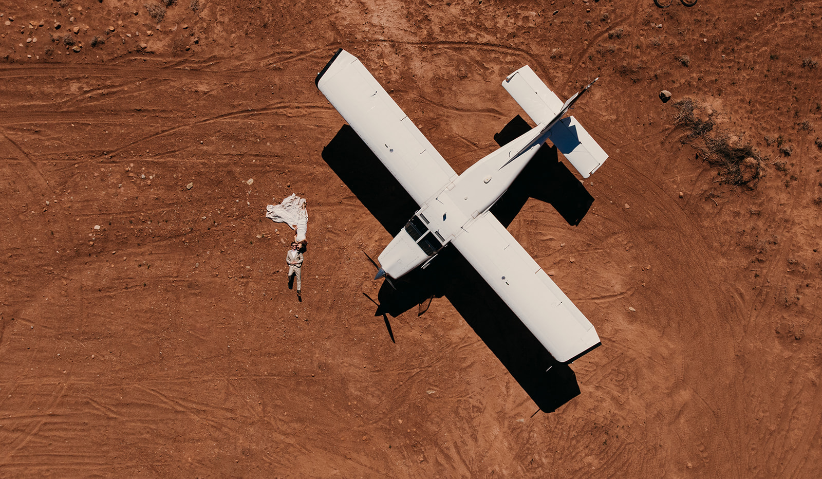 Couple lays on the ground in their wedding attire next to a desert airplane.