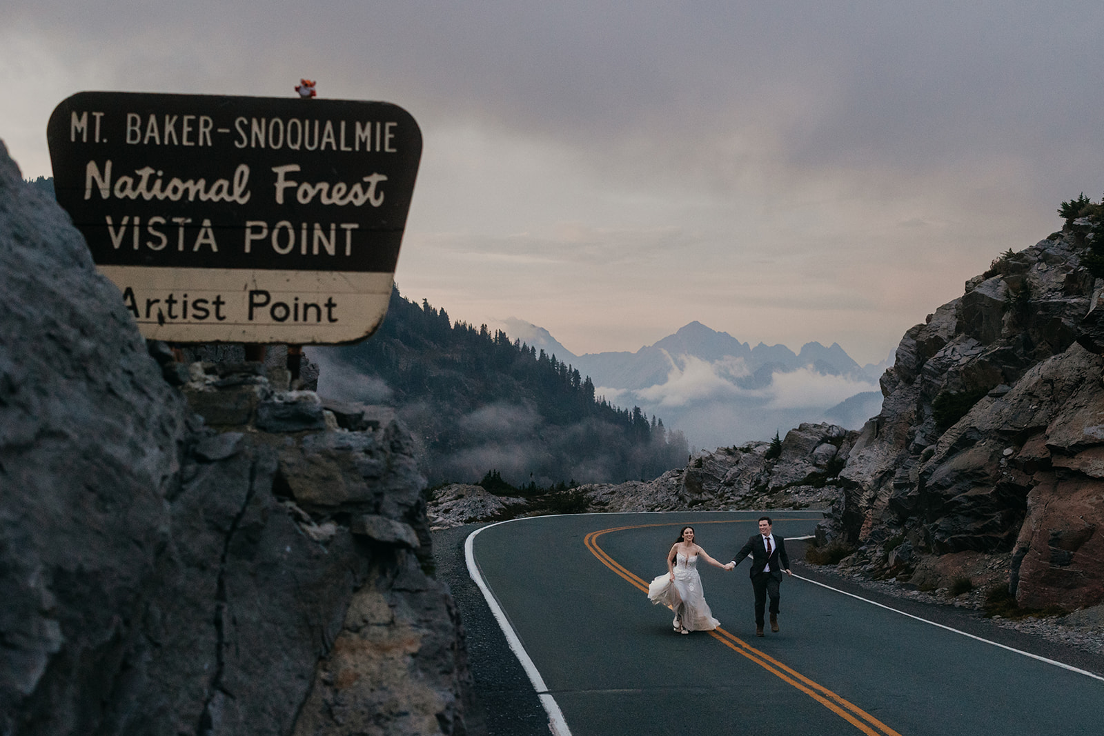 Couple in wedding attire run down the road near Artist Point in Mt Baker-Snoqualmie National Forest.