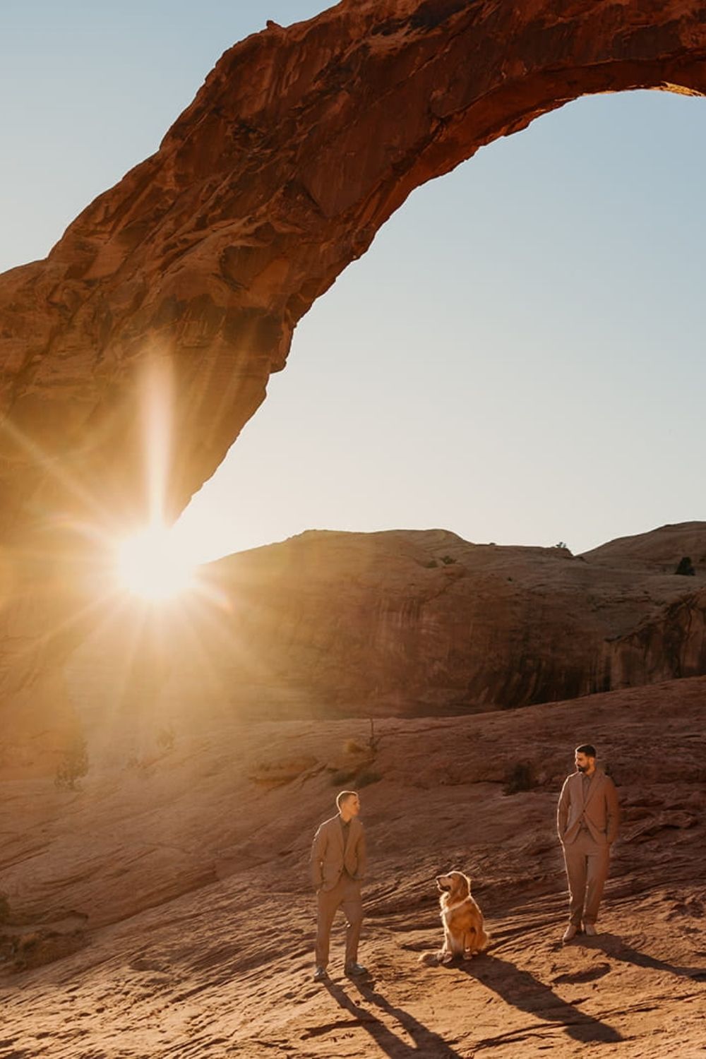 Two grooms stand under Corona Arch with their dog during their elopement at sunset.